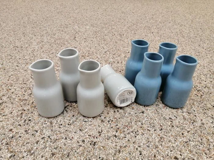 Otte små keramikvaser, fire lysegrå og fire blå, står på en spættet overflade. En grå vase ligger på siden og viser en stregkode på bunden.