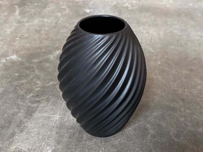 En mat sort, oval vase med en spiralformet, riflet overflade står på en grå betonoverflade.