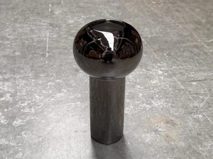 En skinnende metallisk kugle står oven på en cylindrisk metalfod og reflekterer lyset og omgivelserne på en struktureret grå overflade.