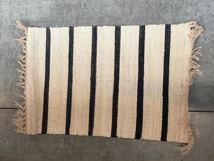 Et rektangulært vævet tæppe med beige og sorte lodrette striber og frynser langs to kanter, der ligger på et gråt betongulv.