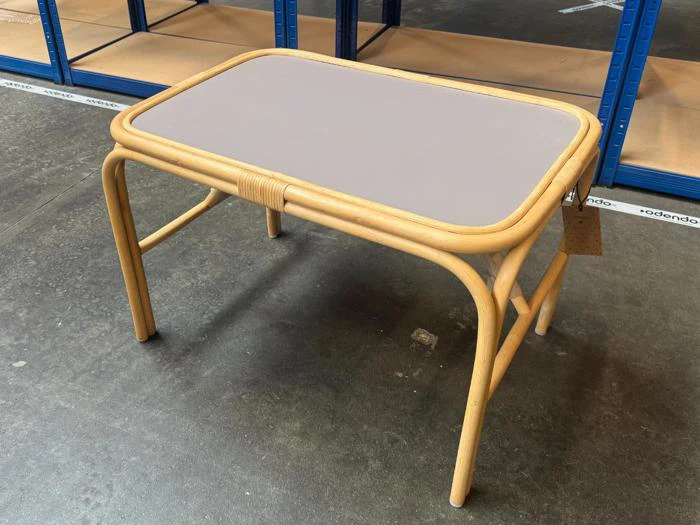 Et lille rektangulært bord med en ramme af lyst træ og en glat, lysegrå bordplade, der står på et betongulv med reoler i baggrunden.