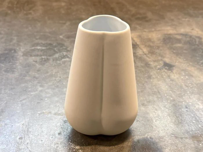 En lille, hvid keramisk vase med en glat, mat overflade og en let uregelmæssig, afrundet form står på en grå, struktureret overflade.