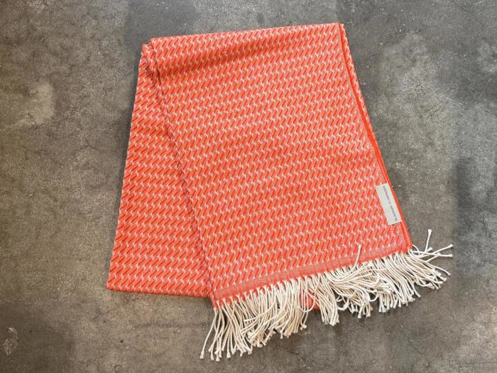Et orange og hvidt vævet tørklæde med frynser i den ene ende ligger udbredt på en betonoverflade.