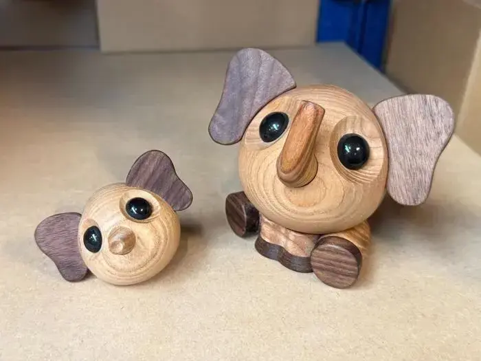 To små elefantfigurer af træ med store sorte øjne og brune ører, hvoraf den ene sidder oprejst og den anden ligger på siden, på en beige overflade.