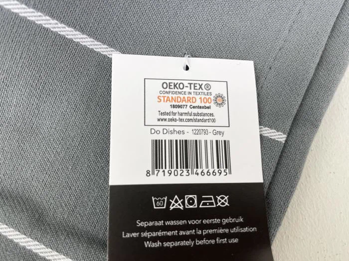 Nærbillede af en tekstilmærkat med OEKO-TEX Standard 100-certificering og produktoplysninger for en grå vare ved navn Do Dishes. Etiketten indeholder også en stregkode og vaskeanvisninger.