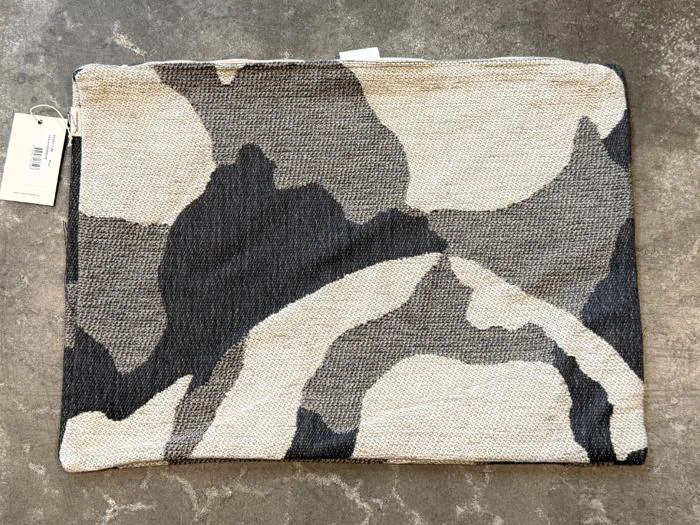 En rektangulær taske med et abstrakt mønster i beige og sort ligger på en struktureret, grå overflade. Der er fastgjort en hvid etiket med tekst til taskenes lynlås.
