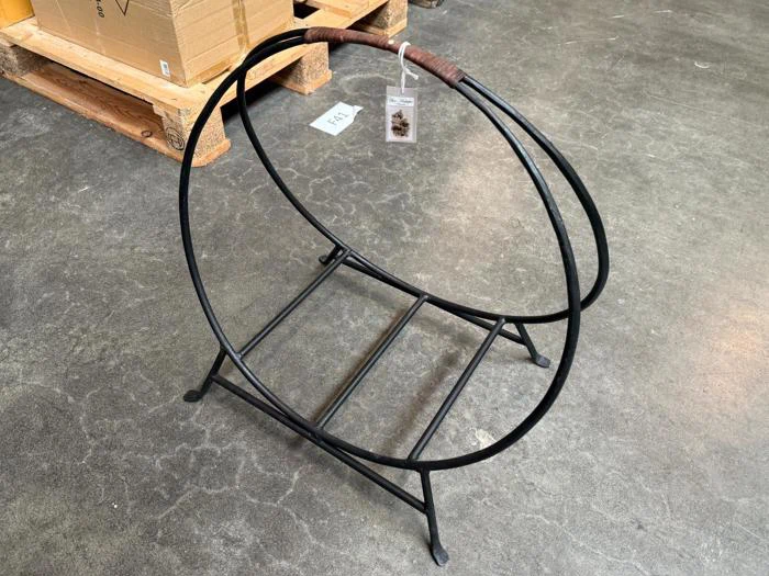 En brændeholder af sort metal med en cirkulær ramme står på et betongulv tæt ved træpaller og papkasser.