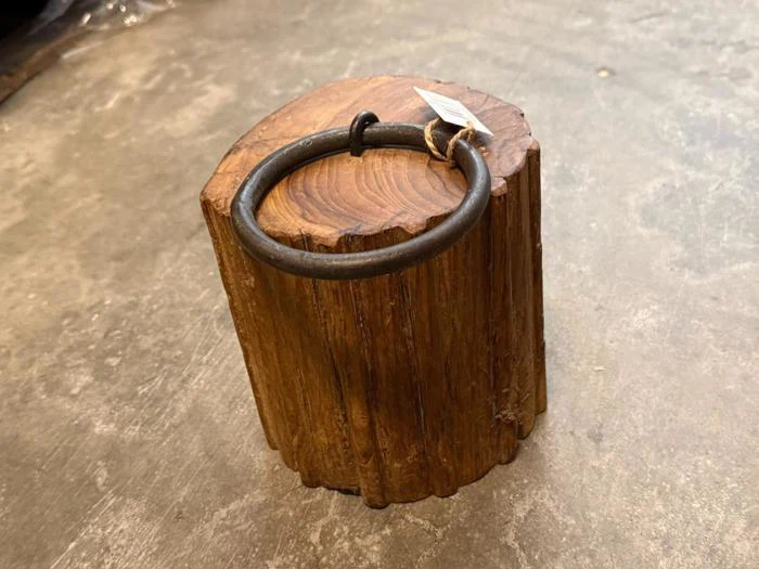 En rund metalring er fastgjort øverst på en lodret træstamme, der står på et betongulv, og der er bundet en lille mærkeseddel til ringen.