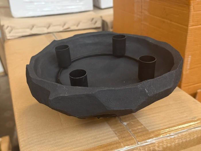 En sort, rund metalholder med struktur og fire opretstående cylindriske sektioner, der står på en papkasse.