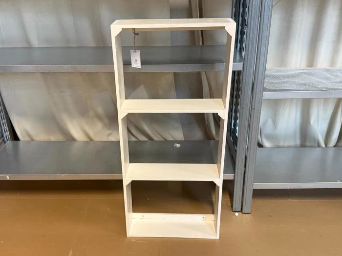 En enkel hvid træreol med fire hylder står på et brunt gulv mellem metalreoler i en butik.