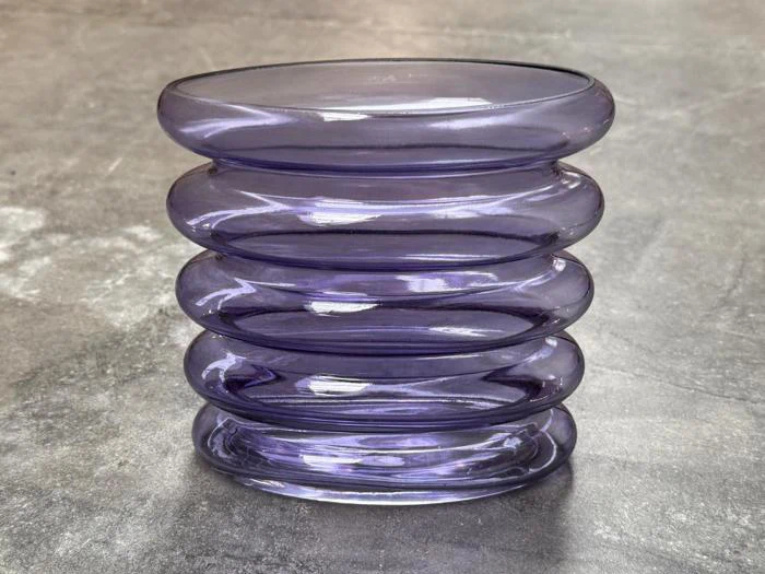 En stak på fem afrundede, lilla glasringe udgør en moderne vase, der står på en grå betonoverflade.