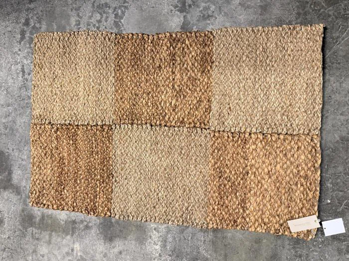 Et rektangulært vævet tæppe med et skakternet mønster i nuancer af beige og brunt, der ligger på et gråt betongulv. Der er fastgjort to mærker i det ene hjørne af tæppet.
