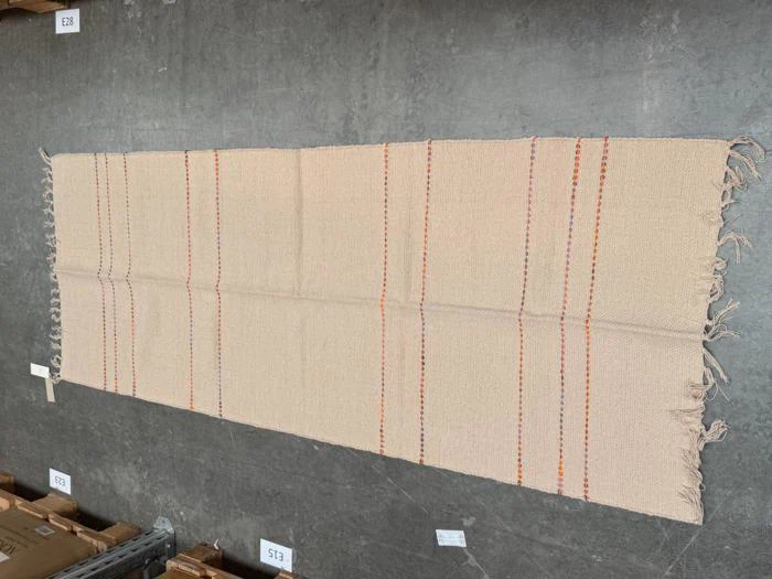 Et rektangulært beige tæppe med tynde orange og lyserøde lodrette striber og frynser langs kanterne ligger på et gråt betongulv.