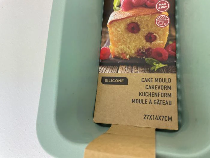 Nærbillede af en silikonekageform med emballage, der viser en hindbærkage og tekst på fire sprog. Målene 27x14x7 cm er synlige.
