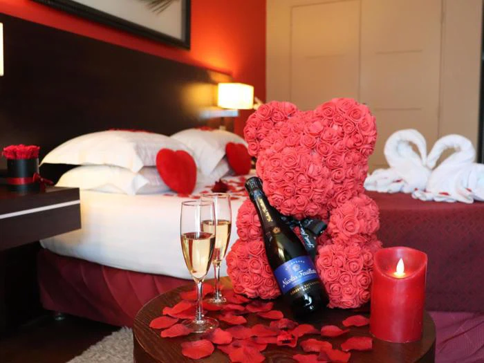Et romantisk hotelværelse med en seng dekoreret med rosenblade og hjertepuder, et bord med champagne, glas, et stearinlys og en bamse lavet af røde roser.