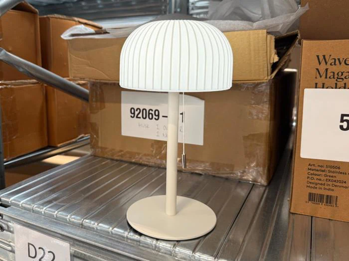 En lille hvid bordlampe med en kuppelformet, riflet lampeskærm står på en metalhylde tæt ved nogle papkasser i en butik eller et lager.