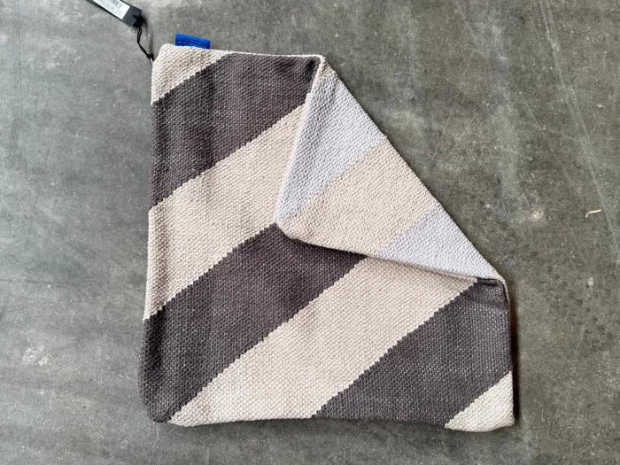 Et foldet, strikket tæppe med brede diagonale striber i sort og beige ligger på en grå betonoverflade.