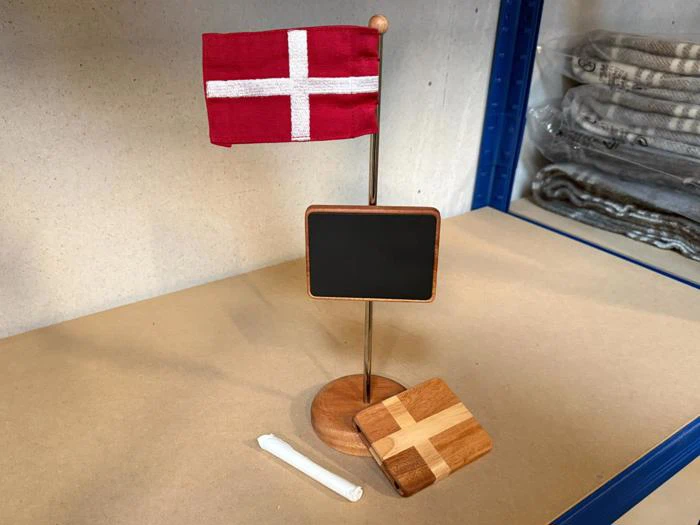 Et lille dansk flag, et tomt tavleskilt, et stykke kridt og en træunderlag med et hvidt kors, alt sammen udstillet på et bord. Sammenfoldede tæpper ligger på en hylde i baggrunden.