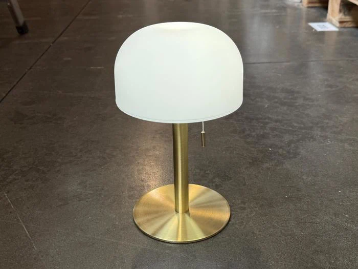 Moderne bordlampe med en rund hvid lampeskærm, messingfod og trækæde, placeret på et gråt betongulv.
