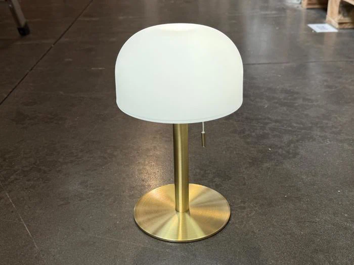 En moderne bordlampe med fod og stamme af messing, kronet af en hvid, kuppelformet lampeskærm, står på et poleret betongulv.