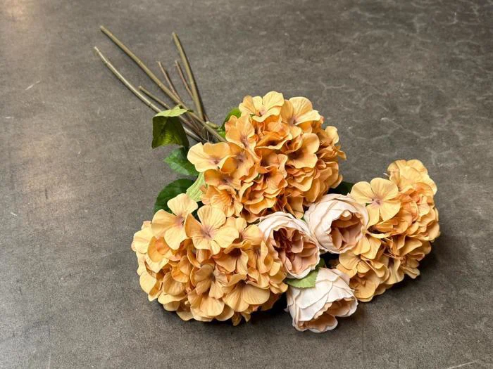 En lille buket af kunstige blomster i farverne fersken og creme med grønne blade ligger på en grå, struktureret overflade.