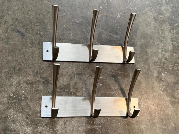 To metalliske vægophængte frakkestativer med tre kroge hver, placeret parallelt på en struktureret, solbeskinnet overflade.