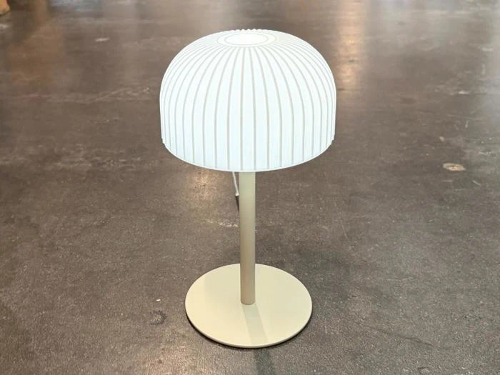 En moderne, hvid bordlampe med en ribbet, kuppelformet lampeskærm står på en rund, beige fod oven på en glat, grå overflade.