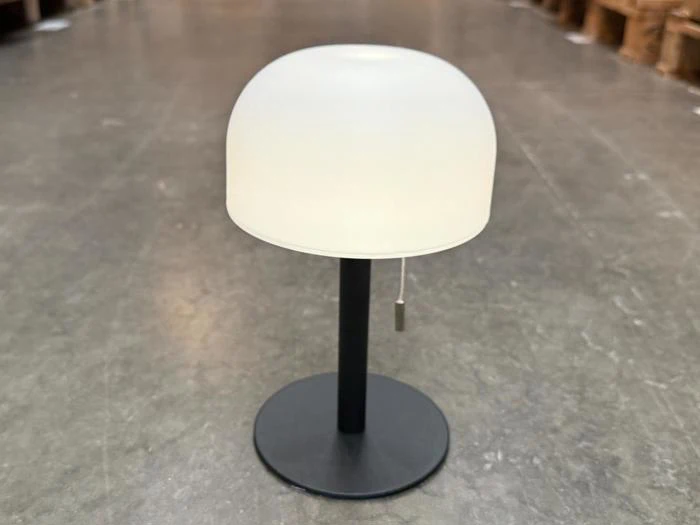 En lille bordlampe med en mat hvid, kuppelformet lampeskærm, sort fod og trækæde, der står på et gråt gulv i et lagerlokale.