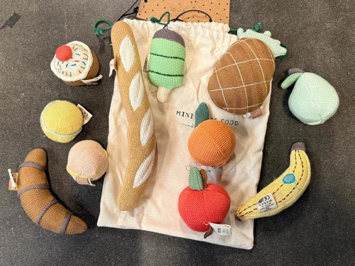 En samling af strikkede legetøjsfødevarer, herunder en baguette, en banan, et æble, en ananas og en cupcake, anbragt på og omkring en opbevaringspose med snor, der er mærket Mini Food.