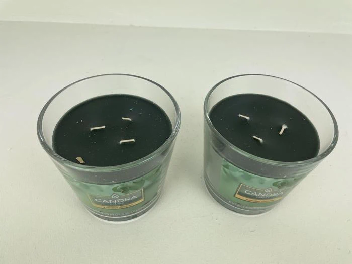 To glaslys med sort voks og tre væger hver, mærket Candra, placeret side om side på en hvid flade.