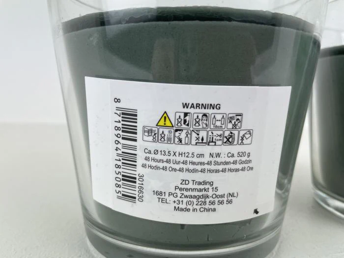 Et nærbillede af et glaslys med en advarselsmærkat med sikkerhedsikoner, produktoplysninger og kontaktoplysninger til producenten.