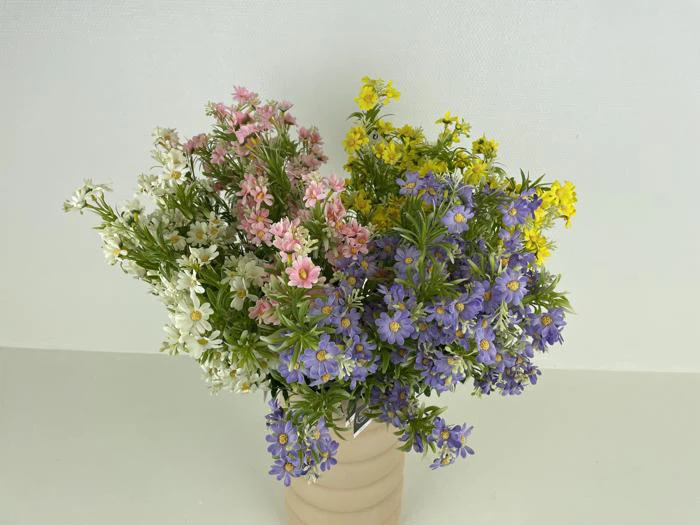 En beige vase indeholder en buket små blomster i hvid, pink, gul og lilla, arrangeret mod en almindelig hvid baggrund.