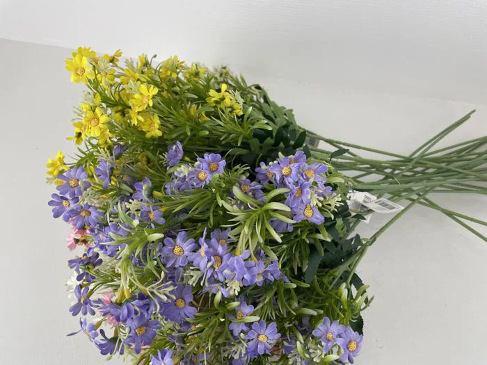 En buket kunstige blomster med lilla, gule og hvide blomster og grønne stilke, der ligger på en hvid overflade.