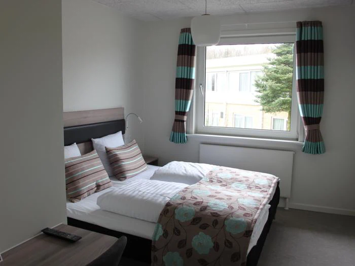 Et pænt hotelværelse med en dobbeltseng, stribet og blomstret sengetøj, et vindue med stribede gardiner, et skrivebord og en fjernbetjening på skrivebordet.