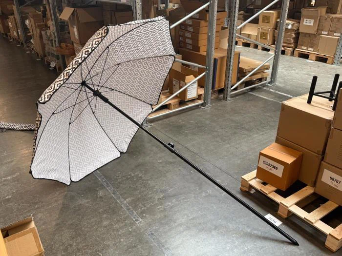 En udfoldet paraply med et sort-hvidt geometrisk mønster på paraplyduken, der ligger på gulvet i et lager nær nogle stablede forsendelseskasser.