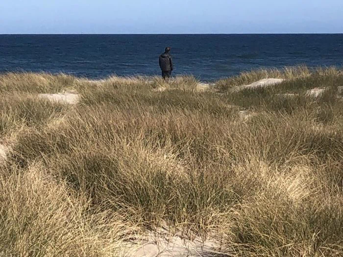 En person står alene på græsklædte klitter med udsigt til det blå hav under en klar himmel.