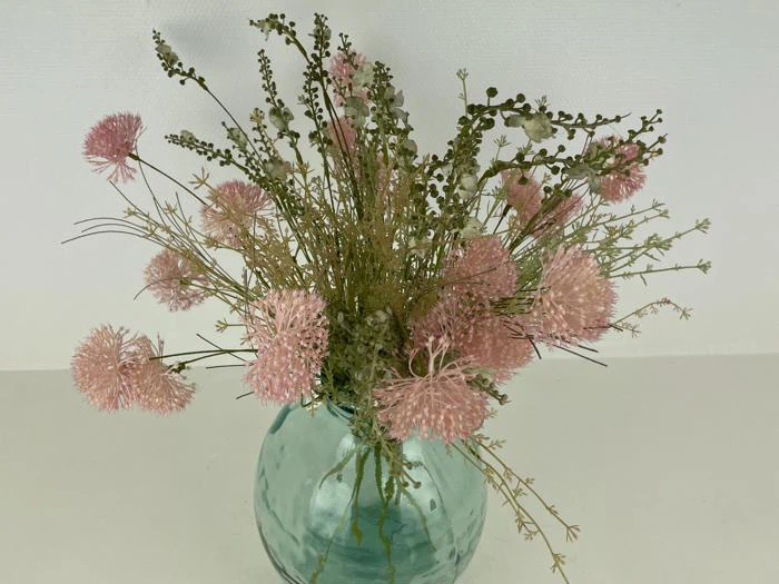 En buket lyserøde og grønne kunstige blomster arrangeret i en rund, lyseblå glasvase på en almindelig hvid baggrund.