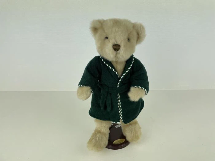 En beige bamse iført en mørkegrøn kappe med hvid kant står oprejst på et lille brunt udstillingsstativ mod en almindelig hvid baggrund.