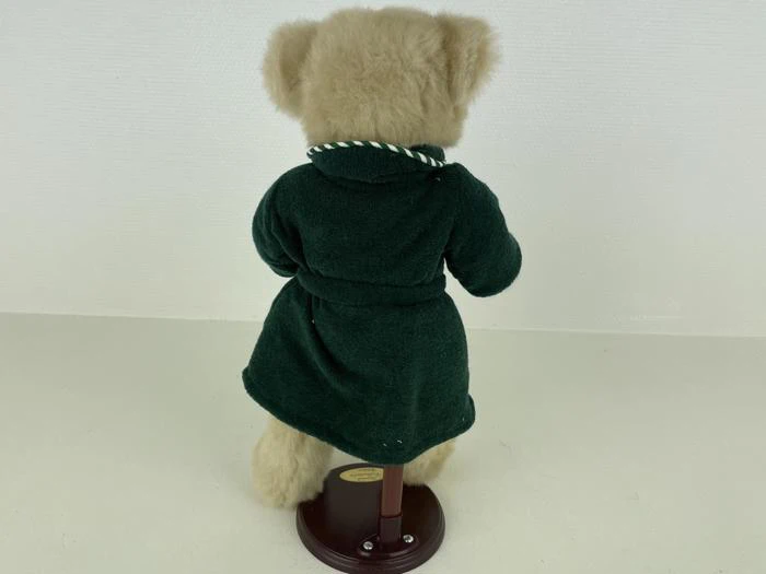 En plysbamse i en mørkegrøn kappe står på et udstillingsstativ og vender væk fra kameraet mod en almindelig hvid baggrund.