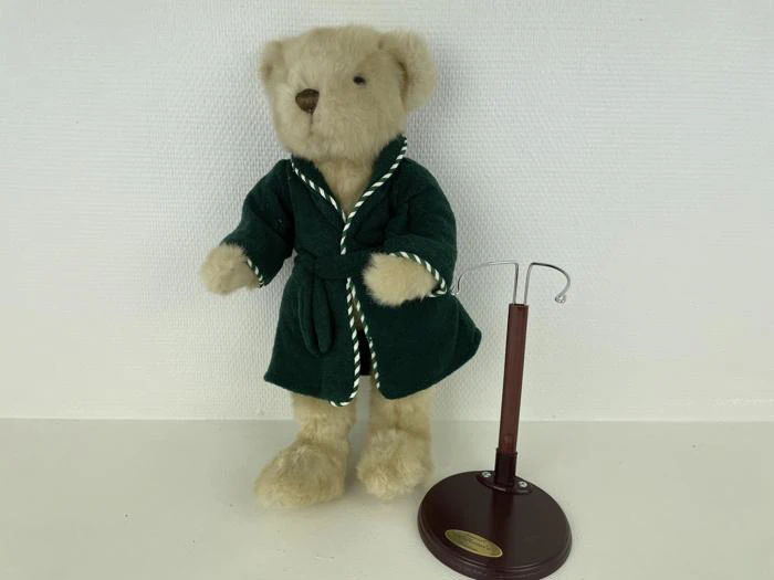 En beige bamse iført en grøn kappe står ved siden af et tomt brunt dukkestativ på en hvid baggrund.