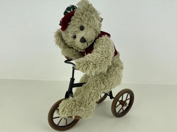 En blød bamse iført rød vest og hue sidder på og krammer en lille sort trehjulet cykel med brune hjul, der poserer på en almindelig hvid baggrund.