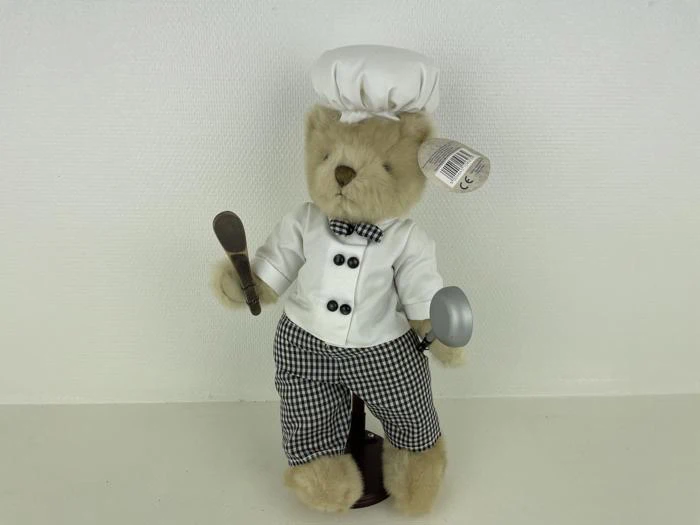 En plysbamse klædt ud som kok med hvid kokkehue, hvid jakke og ternede bukser og med en ske og en grydeske i hånden.