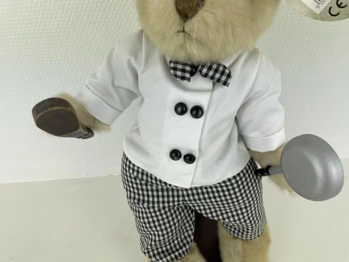 Et udstoppet dyr klædt i hvid skjorte, ternet butterfly, ternede bukser og med en lille grå stegepande i hånden.
