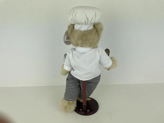 En plysbjørn klædt som kok, iført hvid hat, hvid skjorte og ternede bukser, står på et udstillingsstativ og kigger væk fra kameraet.