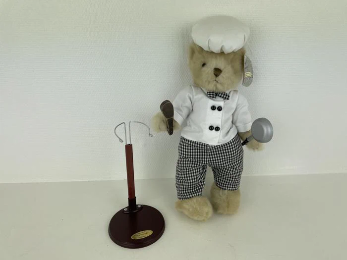 En bamse klædt ud som kok med piskeris og grydeske står ved siden af et rødt og gyldent udstillingsstativ på en almindelig hvid baggrund.