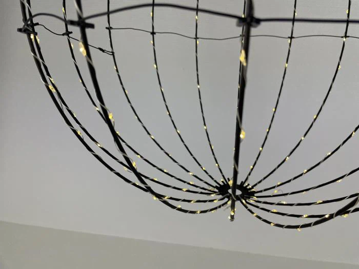 Et nærbillede af en sort trådlampeskærm eller -ramme, formet som en åben kugle, omviklet med små varme hvide lyskæder, mod en almindelig loftsbaggrund.