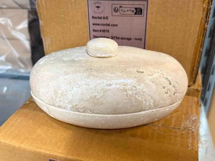 Oval, elfenbensfarvet opbevaringsbeholder af sten med et matchende låg og en knap.