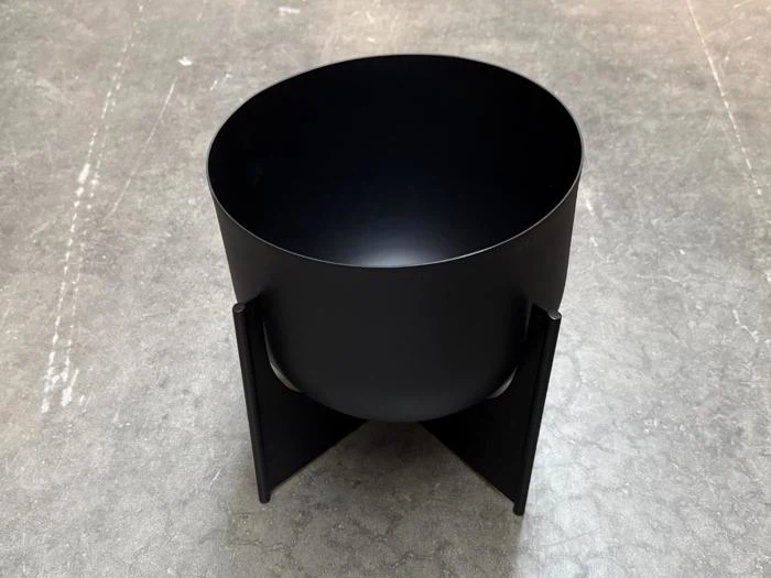En mat sort, cylindrisk blomsterkrukke, der står på et krydsformet metalstativ.
