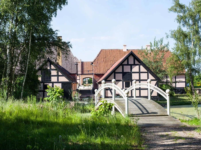 En hvid træbro fører til et traditionelt bindingsværkshus med rødt tegltag, omgivet af grønne træer og græs på en solrig dag.