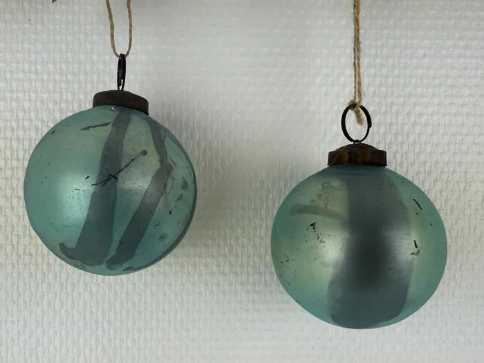 To runde, blågrønne juleornamenter med mørke metaltoppe hænger på snore mod en struktureret hvid baggrund.